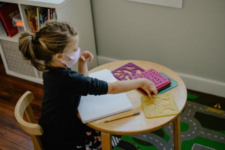 Girl reaching for stencils in a classroom while wearing a mask